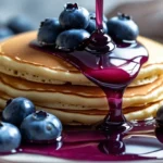 Velvety blueberry syrup drizzling over fluffy pancakes with fresh berries – soft and luxurious breakfast shot