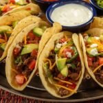 "Family-style taco platter with seasoned ground beef, shredded lettuce, diced tomatoes, avocado slices, shredded cheddar cheese, and fresh cilantro, accompanied by small bowls of salsa, sour cream, and jalapeños on a rustic wooden table."