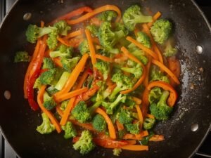 "Broccoli, carrots, and bell peppers being stir-fried in a wok"